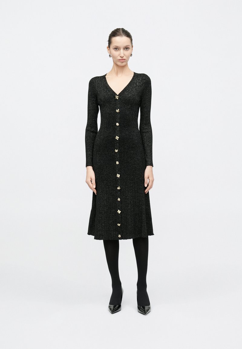 Γυναίκα στέκεται όρθια φορώντας μακρυμάνικο μαύρο πλεκτό φόρεμα με χρυσά κουμπιά μπροστά, μαύρες καλσόν και μαύρα μυτερά παπούτσια.