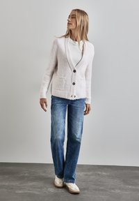 Cardigan di lana rosa chiaro con scollo a V, bottoni frontali, due tasche e orlo a coste, abbinato a jeans blu e sneakers bianche.