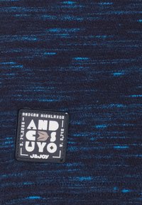 Tissu tricoté bleu foncé avec des éclats de bleu plus clair. Présente une étiquette rectangulaire noire avec du texte blanc et gris indiquant la marque et l'origine.