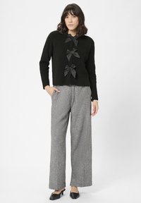 Sort strikket sweater med tre satinbue-detaljer, parret med grå mønstrede vide bukser og sorte slip-on sko.