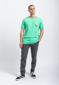 Groene katoenen t-shirt met een klein gedrukt ontwerp op de borst, gecombineerd met grijze elastische broek met enkelbanden en zwarte sneakers.