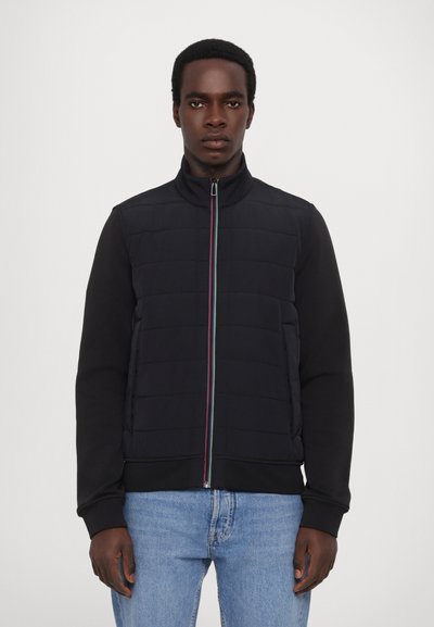 Mørk polstret vest med høy krage, frontglidelås med fargede detaljer, svarte langermede ermer, og ensfarget design; kombinert med lyseblå jeans.