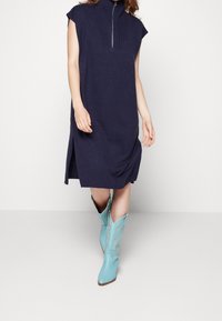Abito a coste blu navy con colletto alto e chiusura con zip. Abbinato a stivali cowboy turchesi con cuciture decorative.