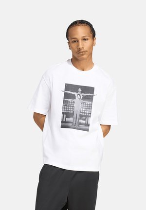 Joven con el cabello trenzado que lleva una camiseta blanca con una imagen en blanco y negro de dos personas de pie con los brazos extendidos.