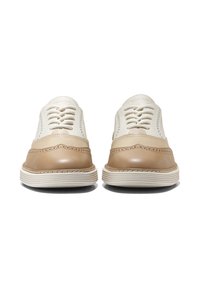 Pár béžových a bílých kožených šněrovacích brogue bot čelících dopředu na bílém pozadí.