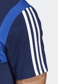 Camiseta deportiva navy con panel azul, que presenta tres rayas blancas en el hombro. Material: tejido transpirable con una textura suave.