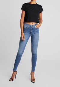 T-shirt nero cropped, jeans skinny a vita alta blu chiaro con sbiaditure e tacchi a stiletto neri. Texture liscia in tutto.