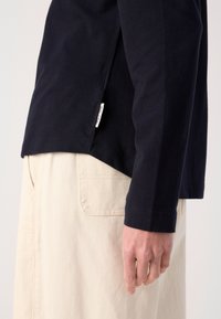 Personne portant un haut noir à manches longues avec une petite étiquette latérale et un pantalon beige clair avec une poche arrière, main reposant le long du corps.