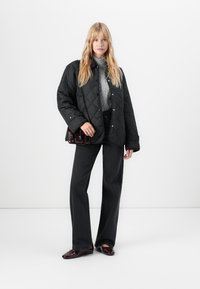 Γυναίκα με μαύρη επενδεδυμένη ζακέτα, γκρι πουλόβερ με λαιμόκοψη, μαύρο παντελόνι φαρδύ, μπορντό τσάντα και αντίστοιχες μπορντό loafers.