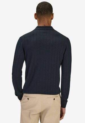 Mand iført en marineblå ribstrikket langærmet sweater og beige bukser, set bagfra mod en ensfarvet baggrund.