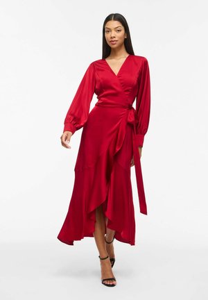 Kvinde iført en rød slå-om-kjole med lange ærmer og flæsekant samt sorte ankelremssandaler, stående mod en ensartet hvid baggrund.