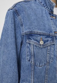 Spijkerjack in lichtblauw met een opstaande kraag, voorzien van twee voorzakken en oranje stiksels. De stof heeft een gestructureerde uitstraling.