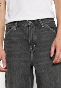 Personne portant un jean noir en denim et un t-shirt blanc, montrant la zone de la taille avec un bouton visible, des passants de ceinture et des poches.
