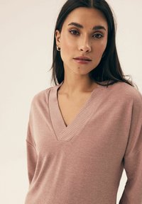 Jasnoróżowy sweter w serek wykonany z miękkiego materiału, z prążkowanymi akcentami przy dekolcie i luźnym krojem. Długie rękawy.