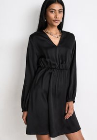 Black dress with a V-neck, long sleeves, and an elastic waist. Made of smooth fabric, it features a relaxed fit and subtle sheen.