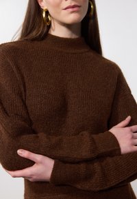 Pull en maille côtelée marron avec col montant et manches ajustées, présentant une texture douce et une coupe décontractée. Aucuns logos ou motifs visibles.