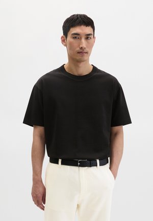 Man draagt een zwart oversized T-shirt in een witte broek met een zwarte riem, staand tegen een effen witte achtergrond.