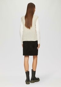 Beige gebreide vest over een witte blouse met lange mouwen, gecombineerd met een zwarte suède rok en zwarte enkellaarsjes met groene zolen.