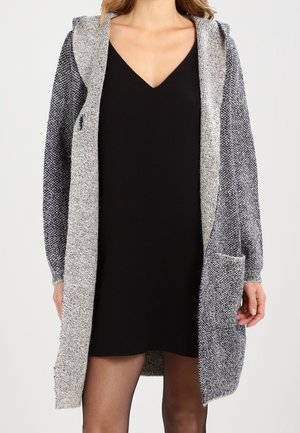 Donna che indossa un vestito nero con scollo a V e un lungo cardigan grigio testurizzato con cappuccio e tasche frontali.