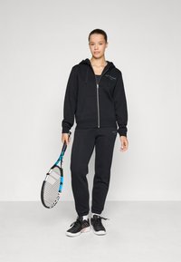 Sweatshirt preto com fecho zip, com bolsos tipo canguru, combinado com calças de moletom a condizer. Modelo segura uma raquete de ténis, vestindo sapatos de desporto pretos com detalhes brancos.
