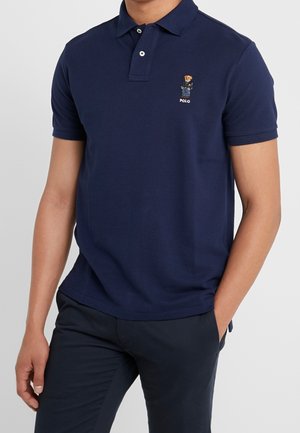 Granatowa koszulka polo wykonana z bawełny, z klasycznym kołnierzykiem, krótkimi rękawami oraz logo misia na lewej piersi. Gładka faktura.