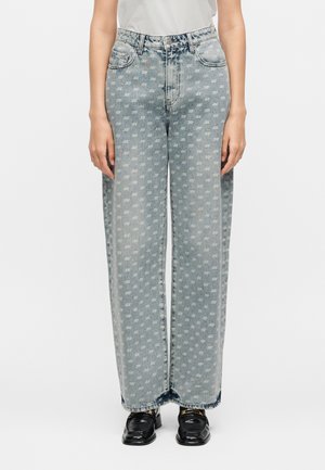Persona che indossa jeans a vita alta e gamba larga color azzurro chiaro con motivo bianco "ax" ripetuto, abbinati a mocassini neri e top bianco.