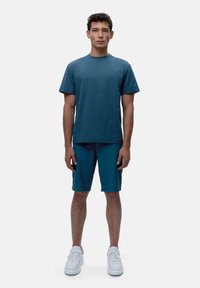 Ciemnoniebieska koszulka z krótkim rękawem w kolorze turkusowym, zestawiona z pasującymi szortami do kolan. Białe trampki o fakturowanej powierzchni dopełniają wygląd.