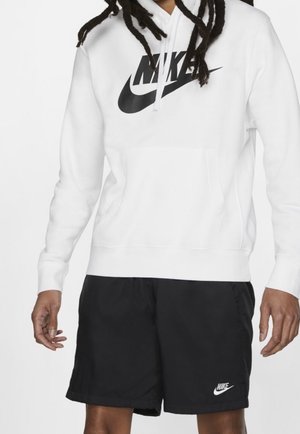 Persona con sudadera con capucha blanca de Nike con logo negro y pantalones cortos negros de Nike con logo blanco, de pie contra un fondo liso.