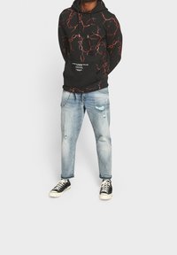 Man in zwarte hoodie met rode bliksempatronen, versleten lichtblauwe jeans met ketting en zwarte lage sneakers met witte veters.
