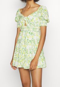 Abito mini floreale in tessuto leggero, con maniche a sbuffo, collo annodato e vita smock. I colori includono motivi floreali verdi e gialli.