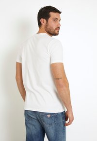 Camiseta blanca de manga corta, con cuello redondo, de tela suave, combinada con vaqueros azules que tienen un logo de triángulo en el bolsillo trasero.