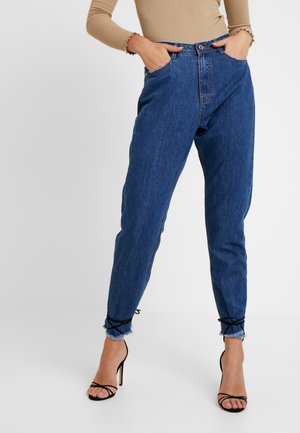 Person trägt blaue High-Waist-Jeans mit ausgefransten Säumen, gebunden mit schwarzen Schnüren, beigefarbenes geripptes Langarmoberteil und schwarze Riemchen-High Heels.