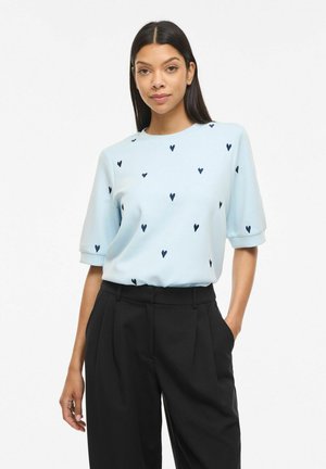 Femme portant un haut bleu clair à manches courtes avec des motifs de cœurs foncés et un pantalon noir taille haute, debout avec une main dans la poche.