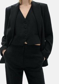 Vrouw gekleed in een op maat gemaakt zwart pak met een geknoopte gilet, blazer en broek, handen in de zakken, tegen een effen lichte achtergrond.