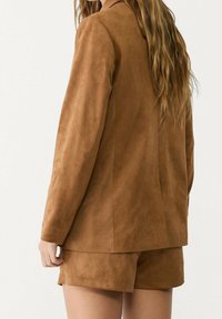 Bruine suède blazer met een getailleerde pasvorm, lange mouwen en een gladde textuur, gecombineerd met bijpassende shorts, die een naadloos ontwerp tonen.