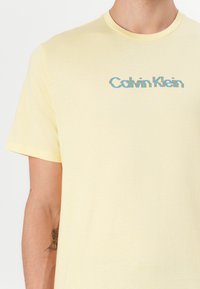 Camiseta de manga corta, color amarillo claro, en una tela suave, con un logo multicolor de "Calvin Klein" en el pecho. Diseño simple y casual.