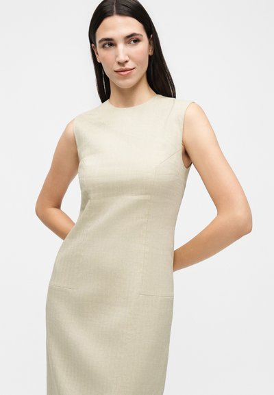 Γυναίκα με μακριά σκούρα μαλλιά φοράει αμάνικο, μπεζ εφαρμοστό φόρεμα μέχρι το γόνατο, στέκεται με τα χέρια πίσω από την πλάτη της μπροστά σε ένα απλό φωτεινό φόντο.