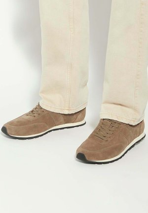 Brown suede sneakers with a black rubber sole, featuring laces and a lightly padded collar, paired with light beige trousers.