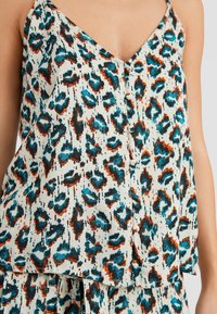 Top sin mangas con estampado de leopardo y escote en V, que presenta patrones en azul, naranja y negro sobre un fondo crema, acentuado por botones.