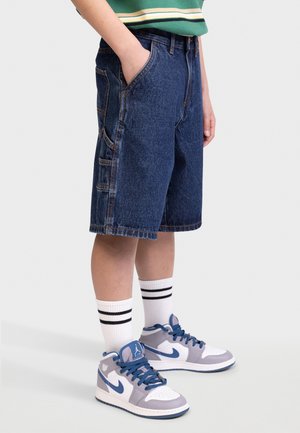 Shorts en denim bleu foncé avec une coupe ample, dotés de poches latérales et de coutures en contraste. Associés à des chaussettes rayées blanches et noires et à des baskets grises.