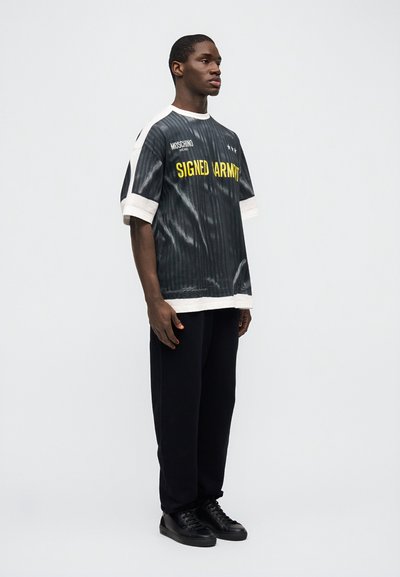 Άνδρας που φοράει μαύρο και άσπρο φαρδύ πουκάμισο με κίτρινο κείμενο, μαύρο  παντελόνι και μαύρα αθλητικά παπούτσια, στέκεται μπροστά σε λευκό φόντο.