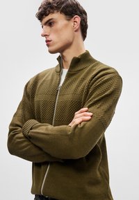 Olive green zip-up sweater with a high collar, featuring textured knitted patterns and ribbed cuffs. The fabric has a smooth finish.