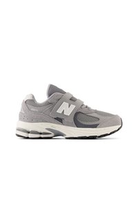 Chaussure de sport grise avec une tige en mesh et des accents en daim, un logo ‘N’ blanc, une semelle épaisse et rembourrée, ainsi qu'une sangle à crochets et boucle.