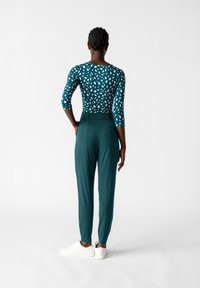 Långärmad, teal top med vitt abstrakt mönster, kombinerad med skräddarsydda, höga gröna byxor och vita sneakers, sett bakifrån.