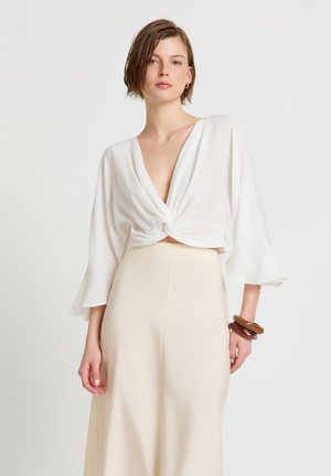 Donna con capelli castani corti che indossa una blusa bianca con scollo profondo a V e dettaglio attorcigliato, gonna beige a vita alta e braccialetti spessi marroni al polso.