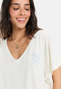 Camiseta blanca, ligera, con cuello en V y mangas cortas. Presenta un logo de palmera azul claro en el lado izquierdo. Textura suave.