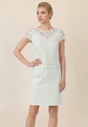 Woman with blonde hair in a white lace short-sleeve top and textured white knee-length skirt, standing with one hand on her hip.