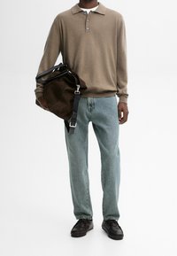 Beige Strickpolo mit drei Knöpfen, hellblauen Akzenten, kombiniert mit hellblauen Jeans und schwarzen Schuhen. Hält eine dunkelbraune Tasche mit einem Schulterriemen.