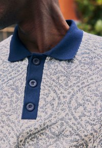 Gros plan sur une personne portant une chemise beige à motif avec un col bleu marine et trois boutons bleu marine sur la patte de boutonnage.