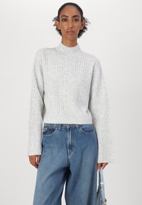 Lys grå strikkegenser med høy hals, ribbestruktur og vide ermer, kombinert med løse blå denimjeans og en liten håndveske.